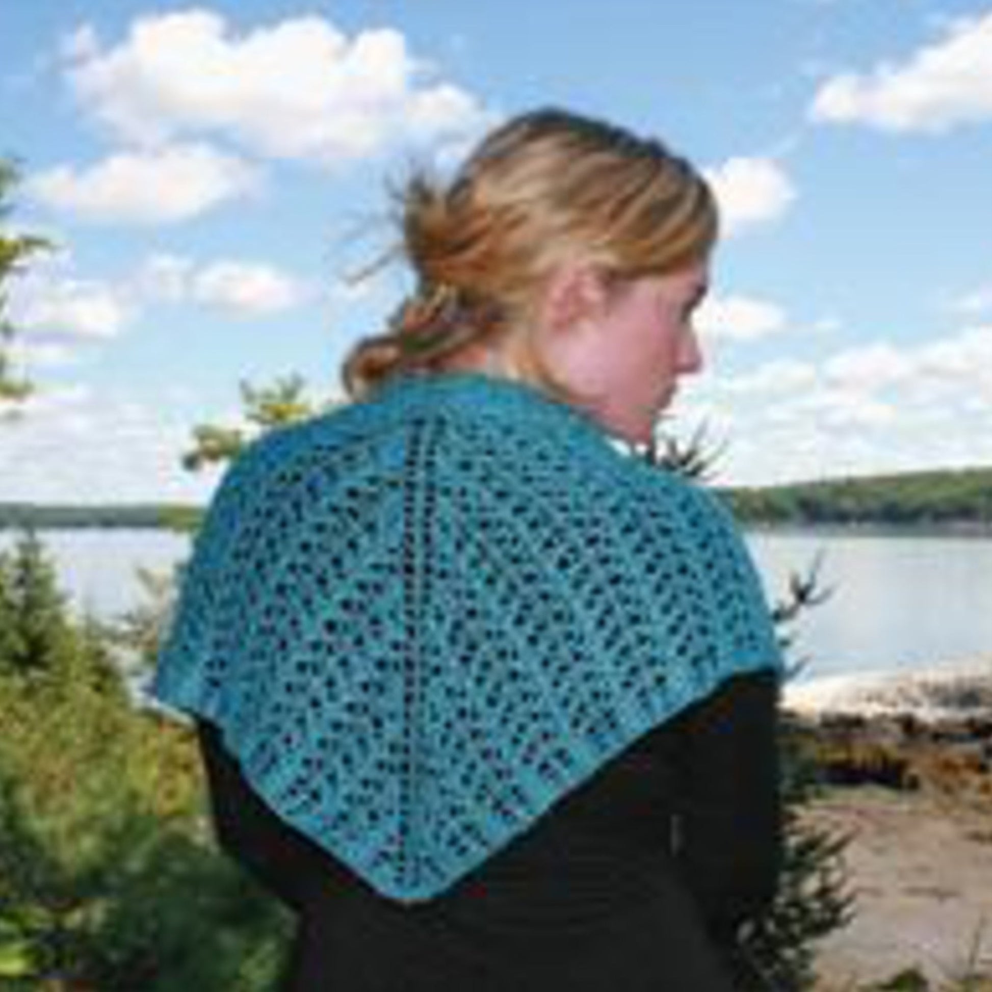 A person with blonde hair tied back is facing away from the camera, wearing the Little Arrowhead Shawl from Halcyon Yarn. The shawl features a beautiful teal color and a scalloped lace pattern, draped elegantly over a black shirt. They are standing outdoors near a body of water, surrounded by trees and a blue sky dotted with scattered clouds in the background.