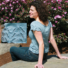 A woman sits on a stone ledge, wearing the Lily of the Valley Shell from Halcyon Yarn. The light blue, short-sleeved knit top features intricate lace detailing with flattering boat neck and scalloped edges. She is surrounded by blooming pink flowers as an inset highlights the elegant lace pattern of her top. Her curly dark hair frames her face, and she completes her look with jeans.