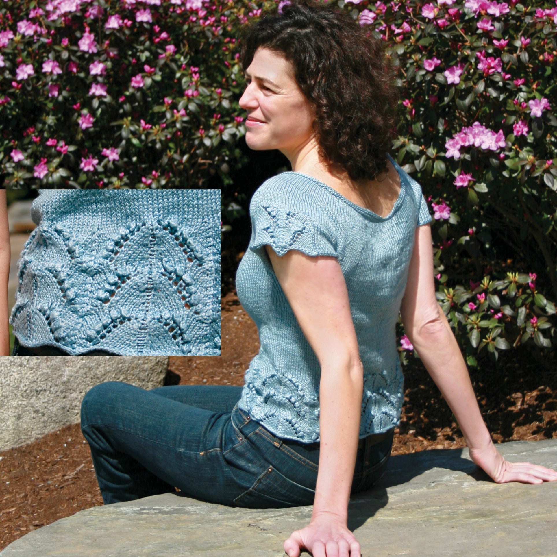 A person with curly hair sits on a stone bench next to a flowering bush, wearing the Lily of the Valley Shell from Halcyon Yarn. The blue, short-sleeved knit top features sweet scalloped edges and an intricate lace pattern. An inset image shows a close-up of the elaborate design on the top's sleeve and hem.