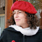 A person with curly hair wears a red Christmas Tree Beret & Mittens from Halcyon Yarn, along with a cream-colored scarf over a black coat. They are looking to the side with a slight smile, with a wooden wall and partially visible window in the background.