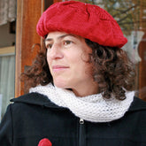 A person with curly brown hair is wearing a Halcyon Yarn Christmas Tree Beret from the Christmas Tree Beret & Mittens set, accompanied by a white knitted scarf. They are dressed in a black coat and looking slightly to the side with a thoughtful expression. The background features a window with a lace curtain and wooden frame.