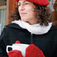 A person with curly hair is donning a red silk Christmas Tree Beret & Mittens from Halcyon Yarn, paired with a white scarf. They are holding a large white cup and wearing a black coat, standing in front of a wooden structure adorned with sheer curtains featuring delicate Christmas tree motifs.