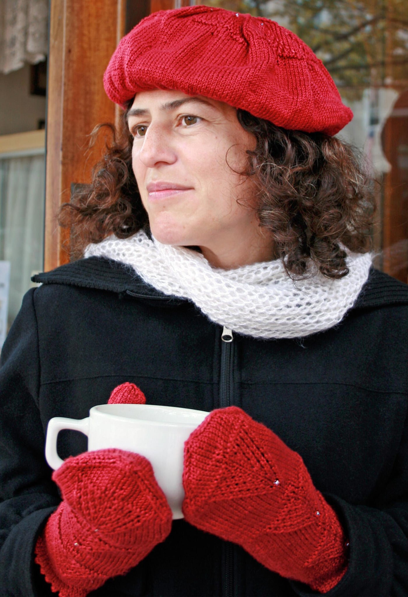 A person with curly hair wearing a festive Halcyon Yarn Christmas Tree Beret & Mittens, a black coat, and a white scarf is holding a large white mug. They are standing outside, looking off into the distance. The background features wooden elements and glass windows adorned with Christmas tree motifs.