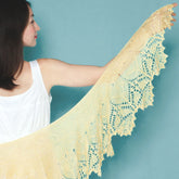 A person in a white sleeveless top stands against a teal background, showcasing the delicate Stellaria Pattern by Twist Collective. Its light yellow fabric features intricate lace borders and forms an elegant crescent shape.