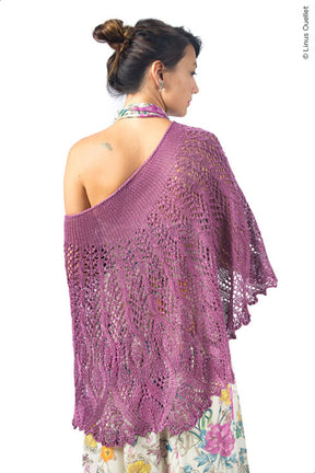 A woman stands with her back to the camera, wearing a beautiful Belarra Shawl by Twist Collective. The shawl features an intricate lace and cable pattern in a stunning shade of purple. Her hair is tied up in a bun, and she is dressed in a floral-patterned dress. The shawl drapes elegantly over her shoulders and down her back, offering a striking contrast to the ornamental grasses swaying nearby.