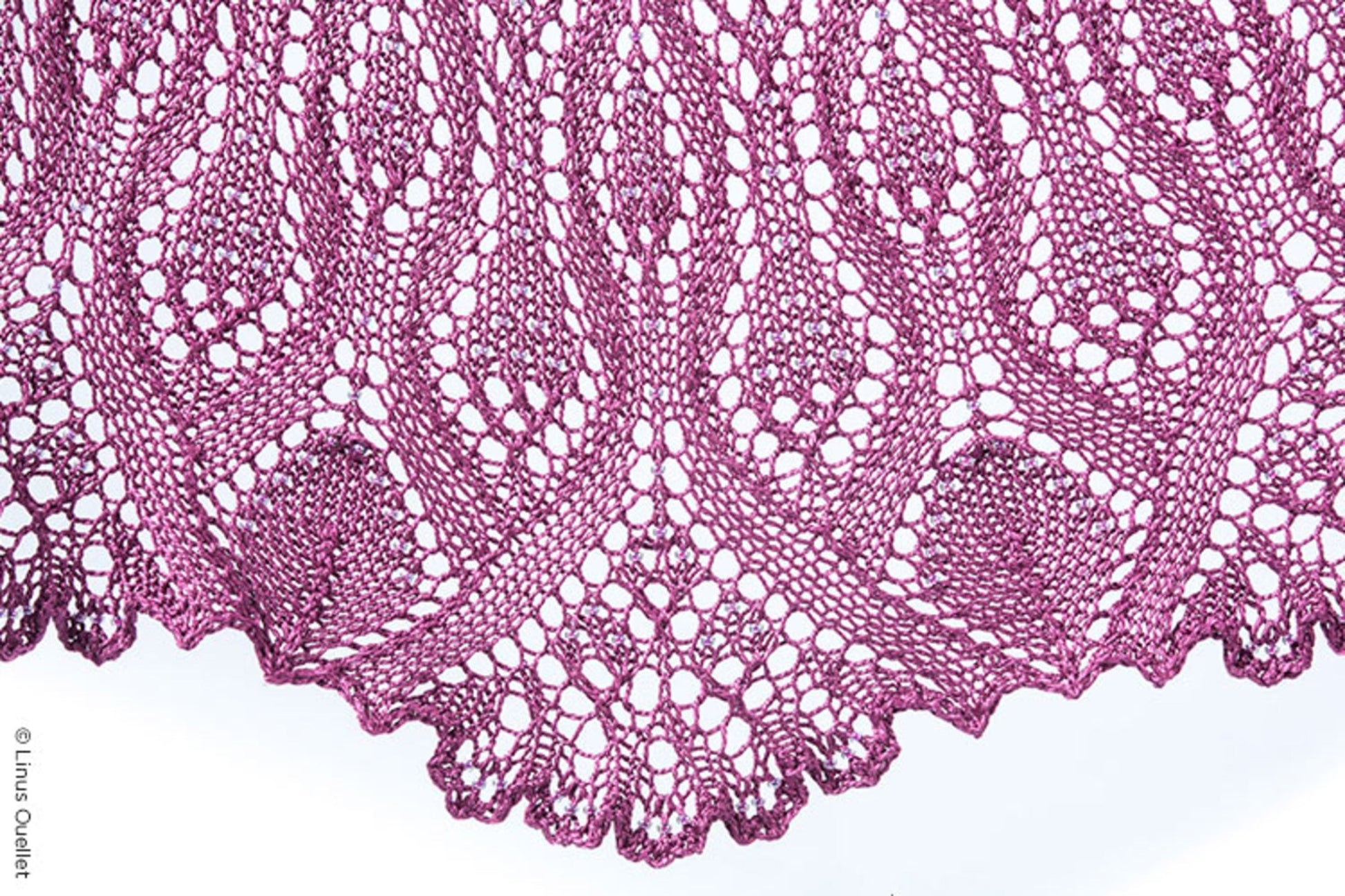 The Belarra Shawl by Twist Collective showcases an intricate knitting pattern crafted from a rich purple yarn. This exquisite design features ornate lacework with repetitive leaf-like motifs and scalloped edges, reminiscent of ornamental grasses. The delicate craftsmanship highlights the fine detail and texture of the knitted fabric.