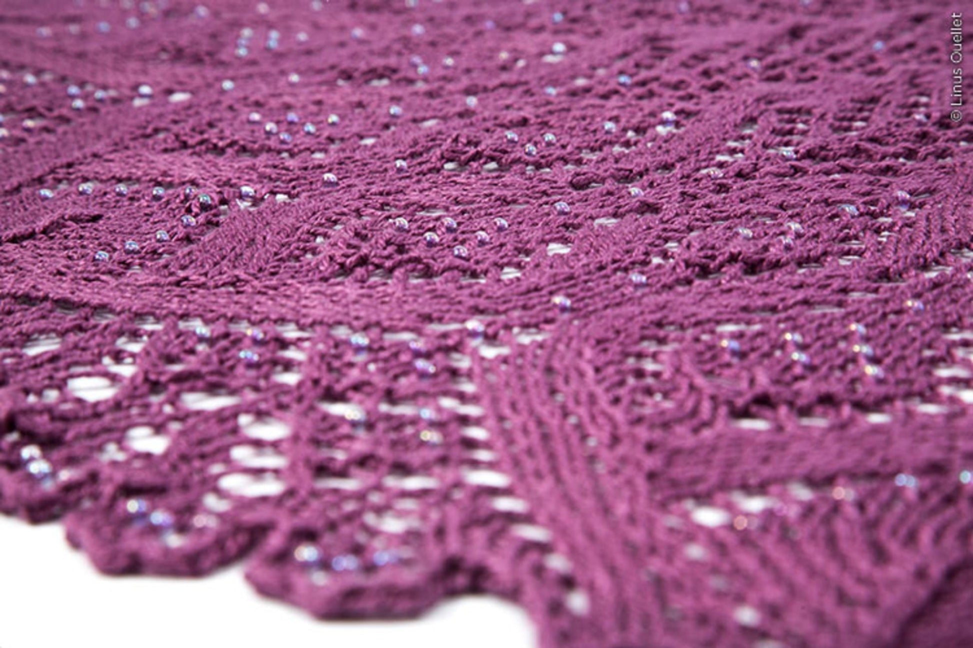 Close-up of the intricately patterned, purple knitted fabric adorned with small, scattered beads. These beads add a subtle sparkle to the textured design, enhancing the overall elegance of Twist Collective's Belarra Shawl. The lace-like edges further contribute to its delicate appearance.