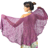 With her back to the camera, a woman gracefully holds out an exquisite Belarra Shawl Pattern by Twist Collective in a rich shade of purple. Her hair is styled in a bun, and she wears a floral dress that perfectly complements the elegant lace and cable pattern of the shawl.