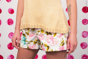 Person modeling a cream-colored Celosia Camisole from Twist Collective paired with floral-printed shorts. Their left arm displays an abstract tattoo. The background features a pattern of large pink polka dots.