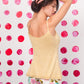 A person stands against a white backdrop with pink and red dot patterns, wearing the Celosia Camisole by Twist Collective, a yellow sleeveless top with crocheted straps, paired with floral shorts. The individual has one hand lifting their hair, revealing a small tattoo on their upper back that appears to be a simple design.