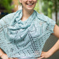 A woman stands outdoors, smiling with her hands on her hips, draped in the Fortuna Shawl—a light blue, intricately knit piece featuring a lace pattern from Twist Collective's Fall 2015 collection. She has dark hair tied back and is in front of a blurred background with green trees and a street.