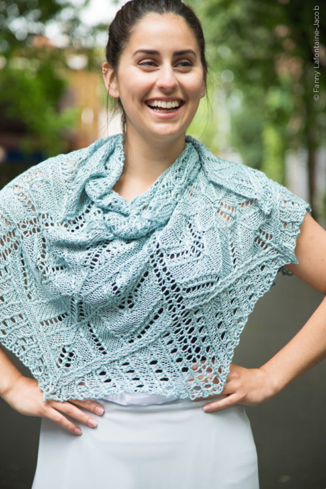 A woman stands outdoors, smiling with her hands on her hips, draped in the Fortuna Shawl—a light blue, intricately knit piece featuring a lace pattern from Twist Collective's Fall 2015 collection. She has dark hair tied back and is in front of a blurred background with green trees and a street.