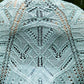 A person is wearing the Fortuna Shawl in light blue, intricately knitted from Gemstone 2/5 Silk. The shawl, by Twist Collective, features detailed geometric and leaf patterns reminiscent of their Fall 2015 collection. Photographed from behind to showcase its full design, the blurry background suggests an outdoor setting.