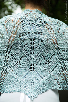 A person is wearing the Fortuna Shawl in light blue, intricately knitted from Gemstone 2/5 Silk. The shawl, by Twist Collective, features detailed geometric and leaf patterns reminiscent of their Fall 2015 collection. Photographed from behind to showcase its full design, the blurry background suggests an outdoor setting.