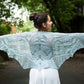 A woman standing outdoors, facing away from the camera, is draped in a large, intricately patterned light blue Fortuna Shawl from Twist Collective. Her arms are outstretched, showcasing the full expanse of the lace pattern in Gemstone 2/5 Silk. She has her dark hair in a bun and is surrounded by greenery.