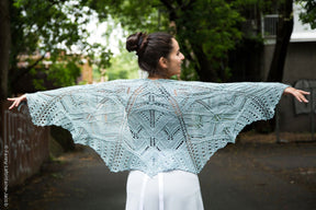 A woman standing outdoors, facing away from the camera, is draped in a large, intricately patterned light blue Fortuna Shawl from Twist Collective. Her arms are outstretched, showcasing the full expanse of the lace pattern in Gemstone 2/5 Silk. She has her dark hair in a bun and is surrounded by greenery.