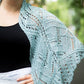 A woman is wearing the Fortuna Shawl in light blue over her shoulders, showcasing the intricate lace pattern popularized by Twist Collective Fall 2015. Dressed in a black top and white skirt against a backdrop of blurred greenery, she smiles with her right hand on her hip, the softness of Gemstone 2/5 Silk evident in the shawl's drape.