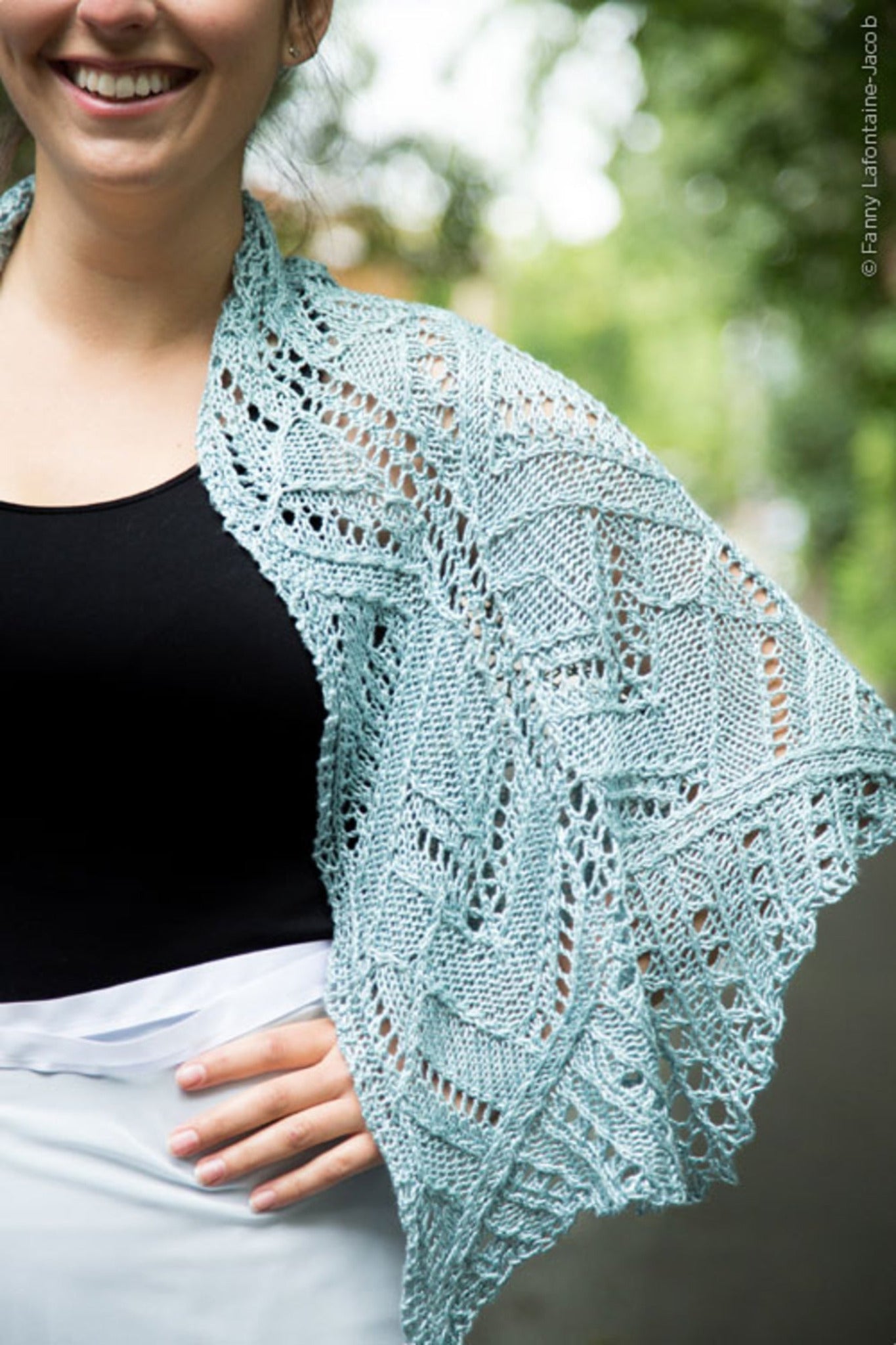 A woman is wearing the Fortuna Shawl in light blue over her shoulders, showcasing the intricate lace pattern popularized by Twist Collective Fall 2015. Dressed in a black top and white skirt against a backdrop of blurred greenery, she smiles with her right hand on her hip, the softness of Gemstone 2/5 Silk evident in the shawl's drape.