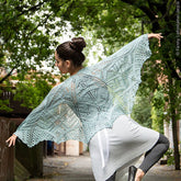 With a bun hairstyle, the person poses gracefully outdoors, draped in a Fortuna Shawl from Twist Collective over a white outfit with gray leggings. They stand on a pathway under lush green trees, reminiscent of an editorial from Twist Collective Fall 2015.