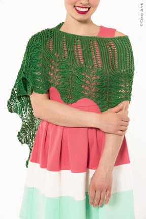 A person wearing a sleeveless dress with pink, white, and green horizontal stripes is posing against a white background. They have the Twist Collective Unnathi Shawl draped over their shoulders, which features an intricate lace pattern. Smiling with their arms folded in front, they exude confidence and style.