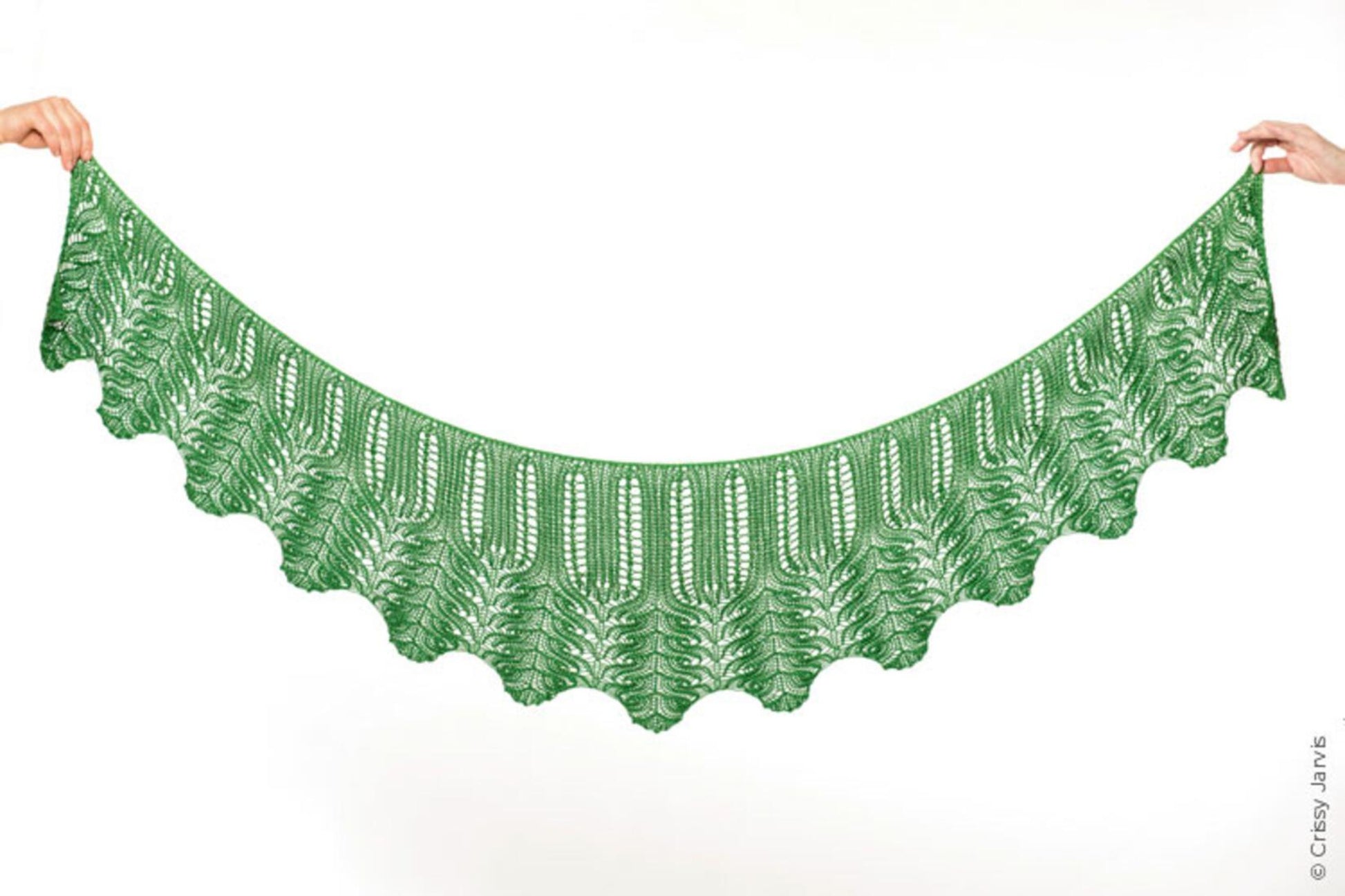 In a plain white background that highlights its exquisite details, a delicately knitted Unnathi Shawl by Twist Collective is held up by two hands at either end. The green shawl showcases intricate lace patterns and a scalloped edge, with an elegant i-cord bind-off.