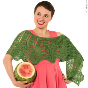 A person with short hair and a bright smile wears the Twist Collective's Unnathi Shawl, featuring a green lace pattern, over a pink dress and holds a halved watermelon under one arm. The background is plain white.