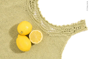 A close-up of a knitted green Papeda Camisole by Twist Collective with a decorative lace yoke. Three lemons rest on top of the camisole, one of which is halved to reveal its insides. The background is white, and © Cristy Johns is visible in the image.