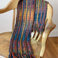 A colorful, warm woven scarf from Halcyon Yarn with a lace ladders pattern, created using a 6-harness weave and soft bulky weight yarn, is draped over a rustic wooden chair. The Lace Ladders Woven Scarf features vibrant hues including blue, red, yellow, and green, with fringed ends. Text on the image reads "Lace Ladders Woven Scarf by Halcyon Yarn - 6-harness weave.