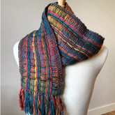 The Lace Ladders Woven Scarf by Halcyon Yarn, made from soft bulky weight yarn, features a textured pattern and stylish fringe at the ends. It is draped around a mannequin's neck, showcasing its gradient color changes in various hues like blues, reds, yellows, and greens arranged in a vertical striped design.