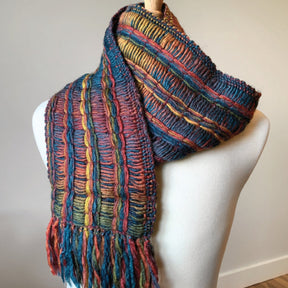 A mannequin displays the Lace Ladders Woven Scarf by Halcyon Yarn, knitted from variegated yarn and showcasing vertical stripes in shades of blue, green, orange, and red. The scarf features a fringe at the bottom and intricate textured patterns running along its length. The background is plain and softly lit, enhancing its winter warmth appeal.