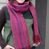 A person is wearing a handwoven, multicolored Chill Chaser Woven Scarf by Halcyon Yarn, featuring shades of pink, purple, and teal with fringe on the ends. They are also dressed in a brown long-sleeve shirt and a green zip-up vest. The person is standing outside next to a gray wall.