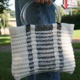 A person holding the Metro Bag by Halcyon Yarn, a handmade knit handbag with clear circular handles. The bag, reminiscent of ribbed bags, features a white and black striped pattern and is held against a backdrop of greenery. The person is wearing blue jeans.