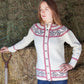 A person with long hair leans against a stack of hay bales in a barn, wearing the Twist Collective's Thornia Cardigan adorned with red and gray patterns. They have a contemplative expression, and a pitchfork rests upright near them.