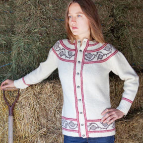 A person with long hair leans against a stack of hay bales in a barn, wearing the Twist Collective's Thornia Cardigan adorned with red and gray patterns. They have a contemplative expression, and a pitchfork rests upright near them.