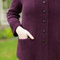 A person dressed in the Sympatico Pattern from Twist Collective stands outside, wearing a purple knit cardigan with black buttons and pockets. The cardigan is perfectly layered over a colorful striped skirt, creating a display of harmonious patterns. One hand rests casually in a pocket against the softly blurred backdrop of a garden.