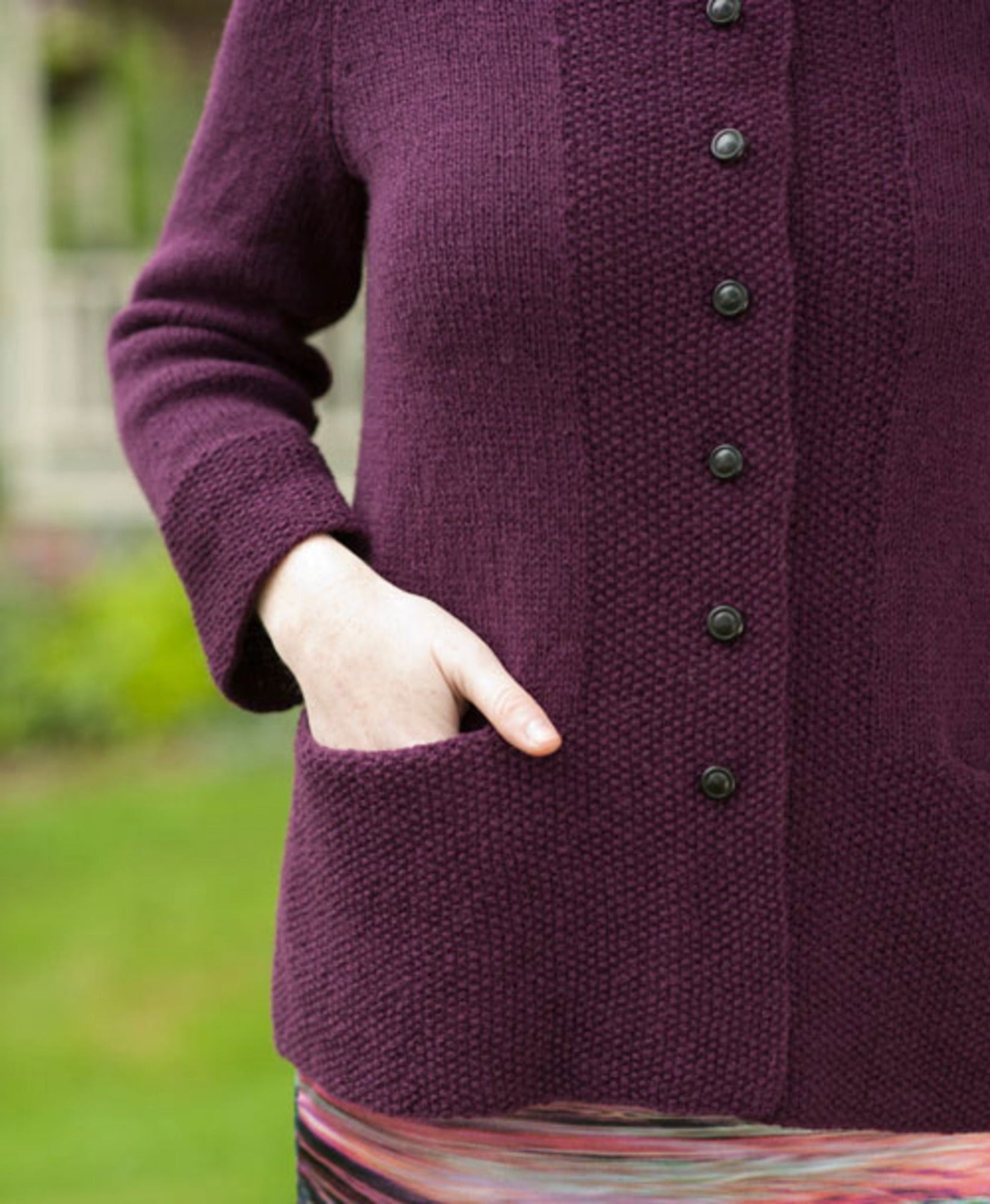 A person dressed in the Sympatico Pattern from Twist Collective stands outside, wearing a purple knit cardigan with black buttons and pockets. The cardigan is perfectly layered over a colorful striped skirt, creating a display of harmonious patterns. One hand rests casually in a pocket against the softly blurred backdrop of a garden.