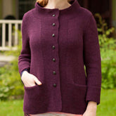 A person standing outside is wearing the Sympatico, a long-sleeved purple cardigan with dark buttons down the front and pockets on either side. The cardigan has a high neckline and features a professional print quality pattern, characteristic of Twist Collective designs. In the background, greenery and part of a house with a porch are visible.