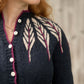 A person with long red hair is wearing the Twist Collective Pinion Cardigan, which features a dark knit design with a white and burgundy feather motif on the shoulders. The cardigan has light-colored buttons and is worn over a necklace with a small circular pendant. The background is blurred.