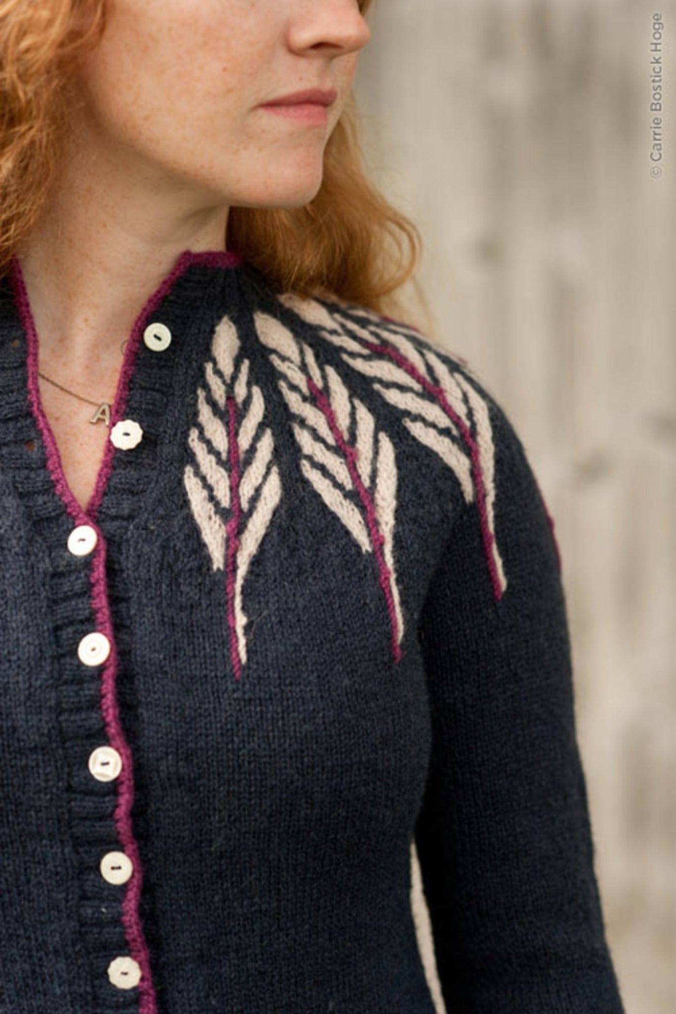 A person with long red hair is wearing the Twist Collective Pinion Cardigan, which features a dark knit design with a white and burgundy feather motif on the shoulders. The cardigan has light-colored buttons and is worn over a necklace with a small circular pendant. The background is blurred.