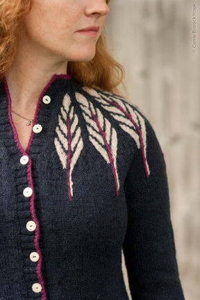 A person with long red hair is wearing the Twist Collective Pinion Cardigan, which features a dark knit design with a white and burgundy feather motif on the shoulders. The cardigan has light-colored buttons and is worn over a necklace with a small circular pendant. The background is blurred.