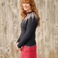 A woman with long red hair is seen smiling and standing in front of a wooden wall in an outdoor setting, wearing the Twist Collective Pinion Cardigan— a black sweater featuring a feather motif on the yoke and raglan shaping—paired with a red plaid skirt. Her hands are relaxed by her sides.