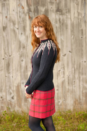 A woman with long red hair is seen smiling and standing in front of a wooden wall in an outdoor setting, wearing the Twist Collective Pinion Cardigan— a black sweater featuring a feather motif on the yoke and raglan shaping—paired with a red plaid skirt. Her hands are relaxed by her sides.