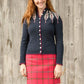 A smiling individual with long, wavy red hair, wearing a Twist Collective Pinion Cardigan adorned with a feather motif on the shoulders and paired with a red plaid skirt, stands in front of a weathered wooden wall. They are also wearing dark tights and have a relaxed, happy expression.