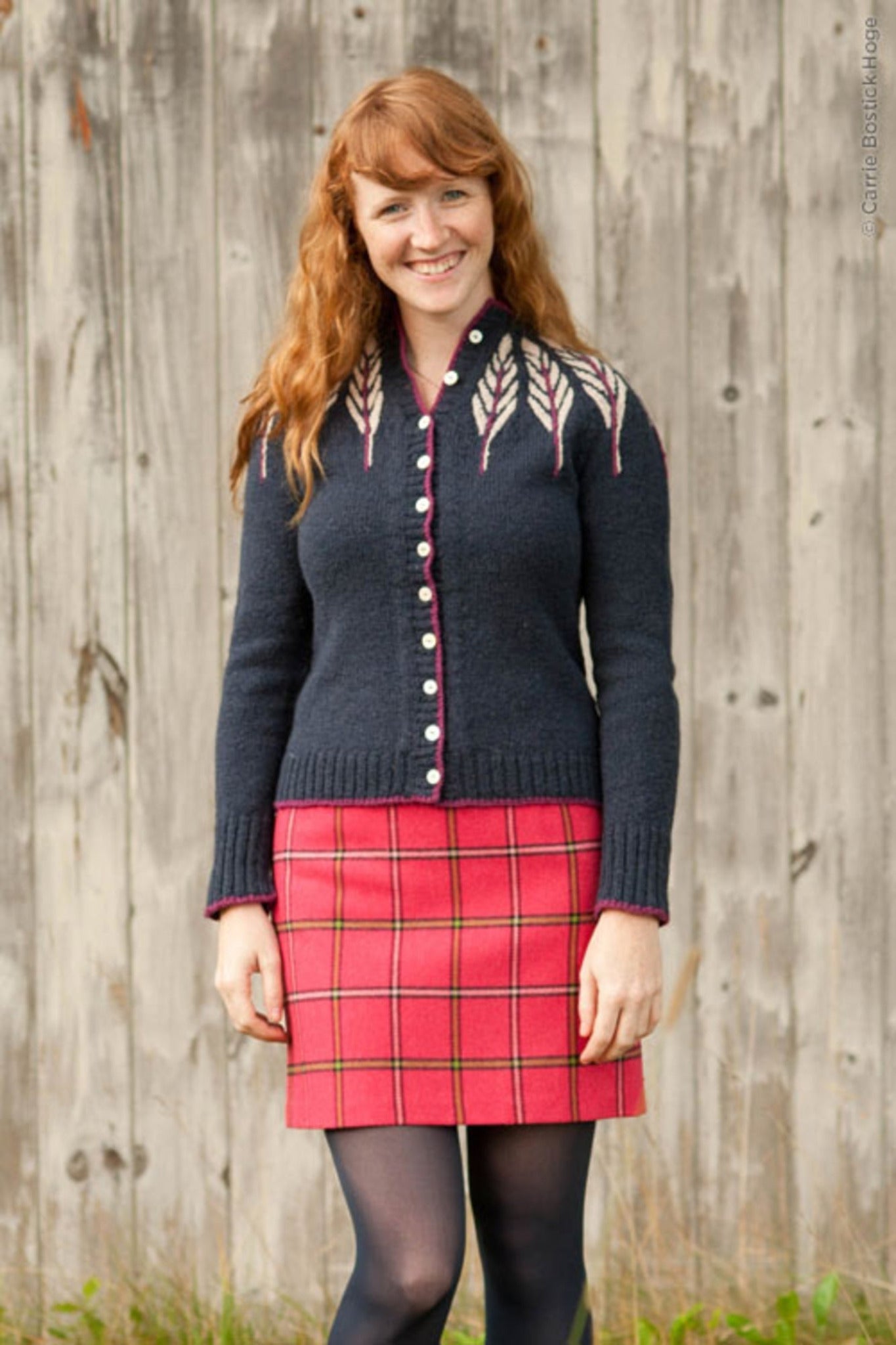 A smiling individual with long, wavy red hair, wearing a Twist Collective Pinion Cardigan adorned with a feather motif on the shoulders and paired with a red plaid skirt, stands in front of a weathered wooden wall. They are also wearing dark tights and have a relaxed, happy expression.