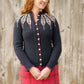 A woman with long, wavy red hair stands in front of a weathered wooden wall. She is wearing the "Pinion Cardigan" by Twist Collective, which features a white leafy feather motif near the shoulders, paired with a red plaid skirt. Looking to the right with a slight smile, her hands are clasped in front of her.