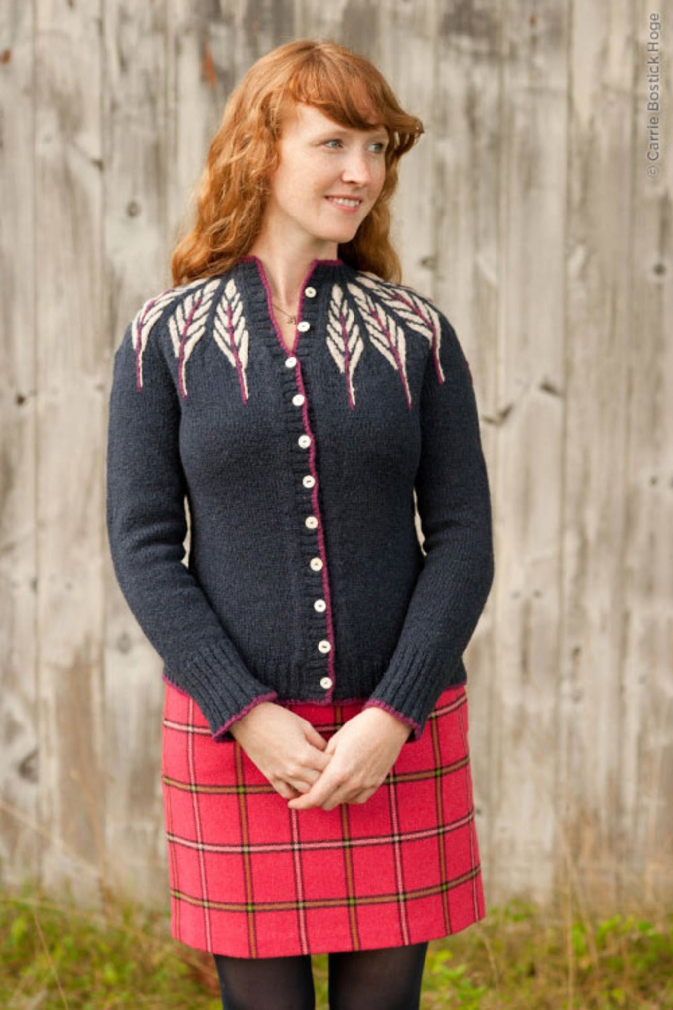 A woman with long, wavy red hair stands in front of a weathered wooden wall. She is wearing the "Pinion Cardigan" by Twist Collective, which features a white leafy feather motif near the shoulders, paired with a red plaid skirt. Looking to the right with a slight smile, her hands are clasped in front of her.