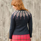 A person with shoulder-length reddish-brown hair is seen from behind, wearing the Twist Collective Pinion Cardigan, a dark sweater adorned with a feather motif pattern of vertical white leaves on the yoke and back. They are paired with a pink and red plaid skirt, standing in front of a weathered wooden wall.