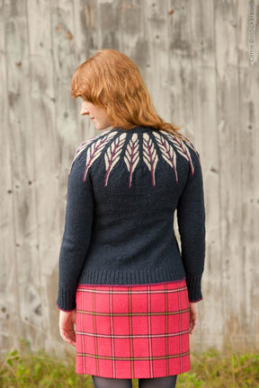 A person with shoulder-length reddish-brown hair is seen from behind, wearing the Twist Collective Pinion Cardigan, a dark sweater adorned with a feather motif pattern of vertical white leaves on the yoke and back. They are paired with a pink and red plaid skirt, standing in front of a weathered wooden wall.