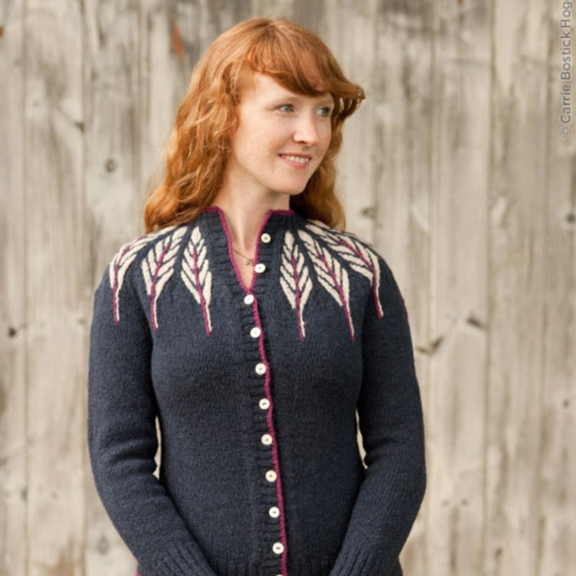 A woman with long red hair is standing in front of a wooden fence and looking to her right. She is wearing the Pinion Cardigan by Twist Collective, a dark blue knitted piece featuring a delicate feather motif around the yoke and a row of buttons down the front.