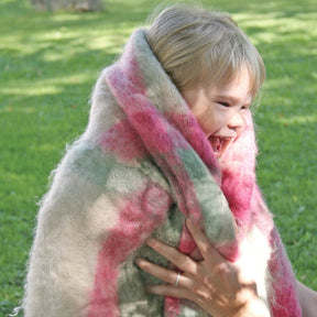 A joyful child, wrapped in a fluffy Victorian Bouclé Spring Garden Woven Blanket from Halcyon Yarn, with shades of pink and green, smiles brightly while a pair of adult hands support them. The background shows a grassy park area in soft focus.